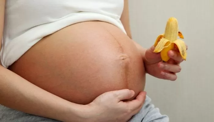 imagem mostrando uma mulher gravida com foco na barriga com a mao embaixo da barriga e a outra mao segurando uma banana pequena